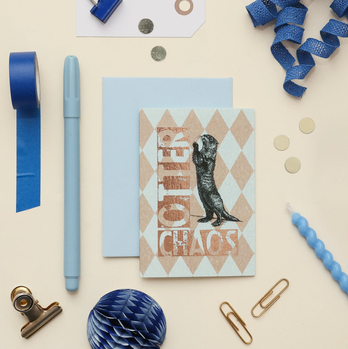 A styled flat-lay of stationery items on a warm cream background, including a light blue greeting card with an otter illustration and the words 'Otter Chaos' in metallic copper lettering, a matching light blue envelope, blue tape, a blue pen, paper clips, coins, and ribbon.