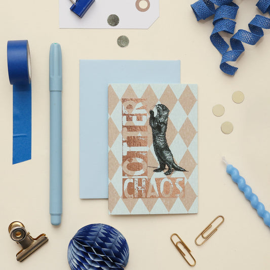 A styled flat-lay of stationery items on a warm cream background, including a light blue greeting card with an otter illustration and the words 'Otter Chaos' in metallic copper lettering, a matching light blue envelope, blue tape, a blue pen, paper clips, coins, and ribbon.